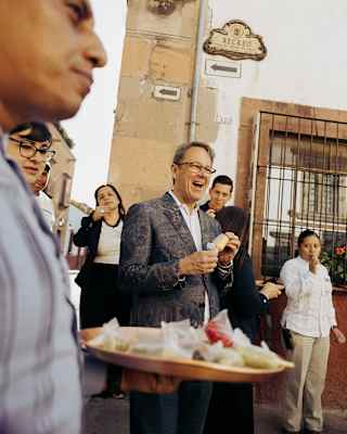 A man in a black suit and glasses unwraps an offering served in the street at a festival, seen over a tray of wrapped sweets.