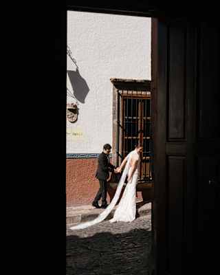 A bride in a white dress and train walks hand in hand with her groom along a cobbled road, viewed from a shadowy doorway.