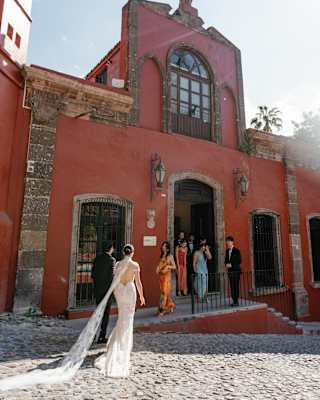 A groom and a bride, trailing a lace train, walk towards guests waiting on the frontage of the terracotta-hued Casa Parque.