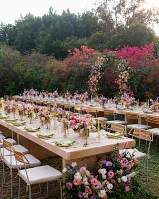 Two long celebration tables, laid with crockery and multiple pink-purple floral centrepieces, wait for guests in the gardens.