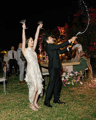 A bride in an ivory slip dress raises two glasses as the groom releases a jet of Champagne from a bottle at an evening party.
