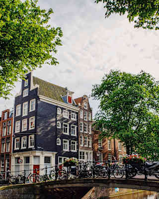 A row of bicycles rests against bridge railings, in a view across a canal that captures tall houses typical of Amsterdam.