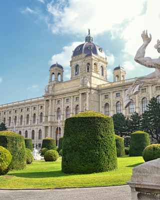 The sandstone Kunsthistorisches Museum, seen past a statue in manicured gardens with topiarised ball and cube hedges.