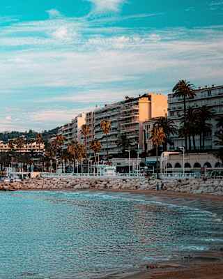 A calm silver-blue sea washes into a bay created by a rock groyne and overlooked by seafront buildings in Cannes.