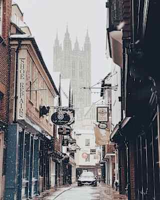Canterbury Cathedral rises in the mist at the end of a narrow street with quaint signage and an approaching Maserati car.