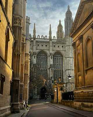 A narrow street leads to a grand Gothic-style building with tall spires and ornate stonework, framed by historic brownstone buildings under a cloudy sky.