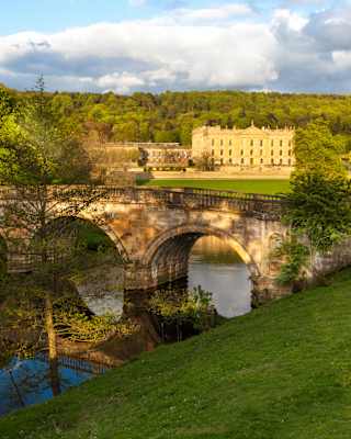 A three-arch stone bridge bows overs the River Derwent as it flows through lush parklands, overlooked by Chatsworth House.