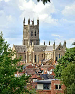 A large Gothic-style cathedral rises above red brick rooftops and trees under a partly cloudy sky. The cathedral features tall, ornate towers and intricate architectural details.