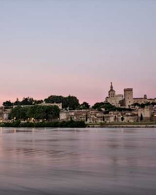 A serene river at sunset with a historic stone bridge on the left and a walled town with a prominent castle or palace on the right, under a pastel pink and purple sky.