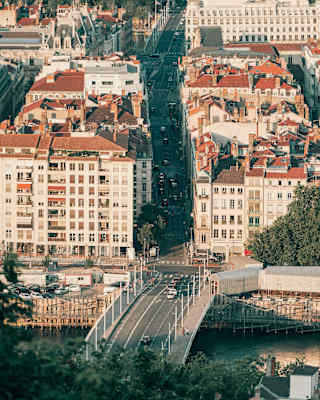 Aerial view of a Lyon bridge over the river Rhone with glimpses of a busy street beyond, shaded by tall buildings either side.