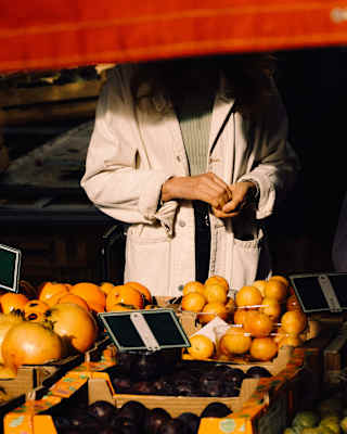 A red awning obscures the face of a man in a white coat who stands at a stall displaying pomegranates, oranges, plums and tomatoes.