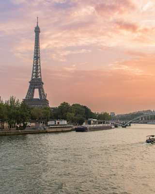 The Eiffel Tower rises against a pink sunset sky in a view across the Seine, where a boat cruises towards a pedestrian bridge.