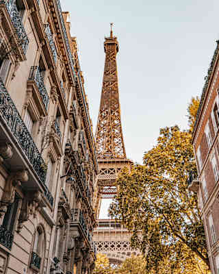 Looking up at the Eiffel Tower from a street flanked by tall buildings, one with iron filigree Juliet balcony railings.