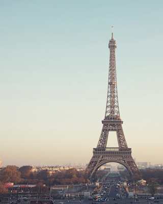Atmospheric image of the Eiffel Tower straddling a road and rising into a dusky blue sky as sunshine brushes distance roofs.