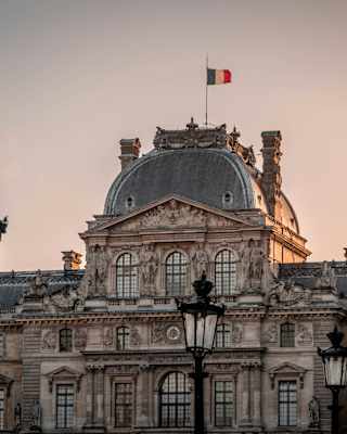 A French flag flies atop the Louvre's Pavilion Sully in a view of the museum at sunset, with gold sun and purpling sky.