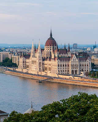 The image shows the Hungarian Parliament Building on the bank of the Danube River in Budapest, with surrounding city buildings and hills in the background under a partly cloudy sky.