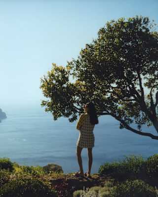 A woman in a short dress with long dark hair stands on a hilltop by a tree, taking in the spectacular Amalfi coast views.