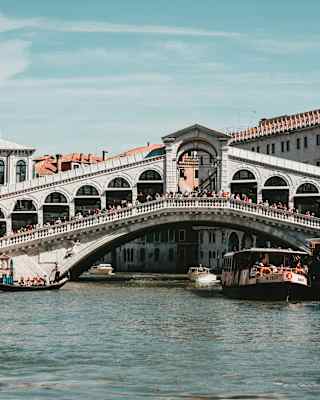 Crowds bustle across the Rialto Bridge in a view from the canal, capturing blue sky above and pleasure boats either side.