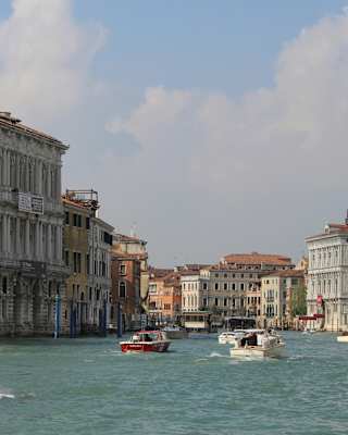 Focus is on wooden boat in the foreground, in front of several others cruising the Grand Canal on a sunny day, seen at eye-level.
