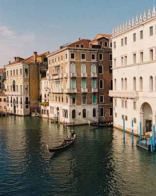 A gondola cruises the Grand Canal towards a side passage where another boat emerges between the rio's historic buildings.
