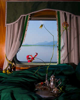 A tray with a silver tea set rests on a green duvet adorned with a flame lily that stands out against the misty window view.