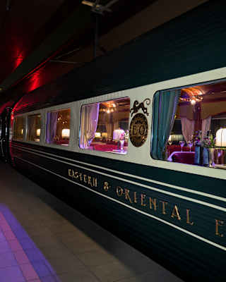 Lights glow in the restaurant car as the train waits on a platform with red and purple lights, seen at night from outside.