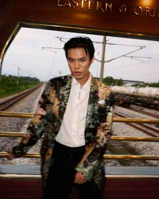 Grainy image of a man in a patterned blazer in the Observation Car with tracks and wires behind, photographed by Kitajima.