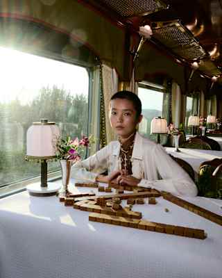 A female model with short dark hair and a white top sits at a clothed table with a lamp and scattered game blocks.