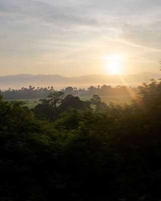 Soft sun glows in a part-cloudy sky, rising above misty, silhouetted hills, seen from the train over fields and trees.