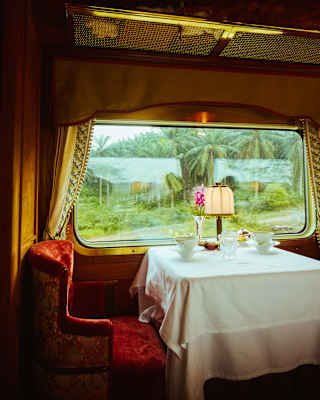 A table set for two with a white tablecloth, lamp, and flowers sits by a large window in a vintage railway carriage, with greenery visible outside.