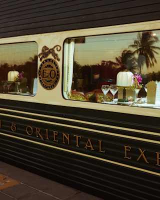 A close-up of the Eastern & Oriental Express train shows a window with a dining table set with glasses and plates; palm trees are reflected in the glass.