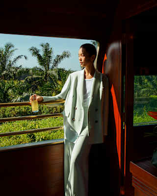 A woman with short dark hair in a loose, mint trouser suit stands on the part-shaded Observation Car deck, holding a drink.