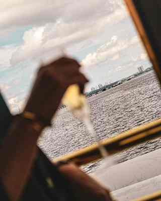 Tilted shot of a body of water seen through a train window, part-obscured by hand holding a Champagne glass in soft-focus.
