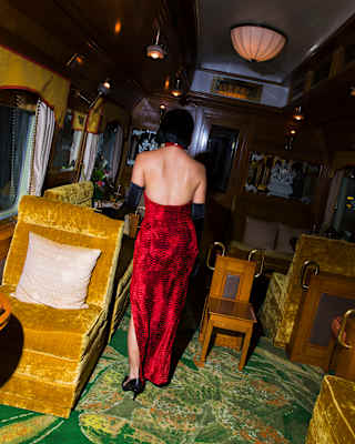 A woman in a red, backless dress stands in the Piano Bar with one gloved hand on a gold-tone velvet seat, seen from behind.