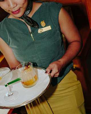 A waitress in a green sleeveless top and mustard skirt carries a tray with a clear-spirit cocktail and a tumbler with bitters hues.