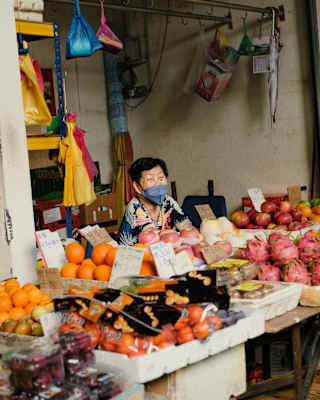 An older woman wearing a hygiene mask sits behind her fruit stall of oranges, apples and dragon fruits in a local Penang market.