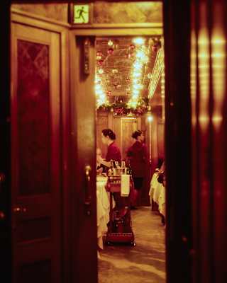 View through a doorway into a dining car where two women walk between tables beneath strips of warm-glowing ceiling lights.