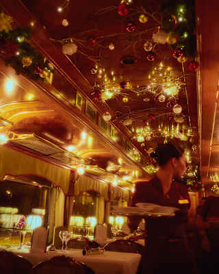 Red and gold baubles hang from the ceiling of a dining car glowing with warm lamps, where a waitress walks between tables.