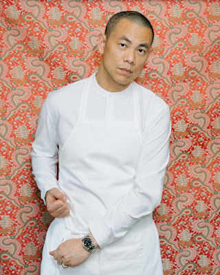 Chef Andre Chiang stands against a red and green-patterned wall, tightening his white apron straps while looking to camera.