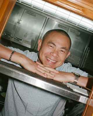 Tilted image of Chef André Chiang, smiling as he rests his chin on his hands, which rest on a steel shelf at the kitchen pass.