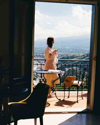 A woman in a robe stands at her balcony railings drinking coffee as she gazes at Taormina's hills, seen from inside a suite.