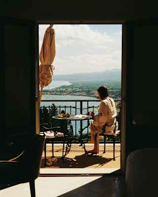 A dark-haired woman reads a magazine at her balcony table, overlooking Taormina's coastline, seen from inside the suite.