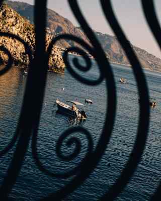 A boat floats on calm sea near a rocky headland, with others further out, glimpsed through curled wrought iron railings.