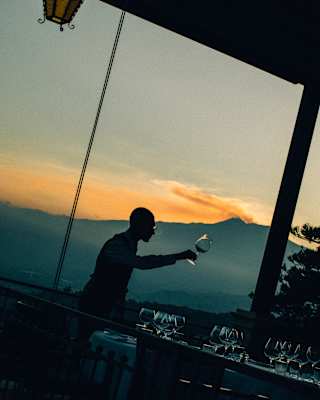 Angled image of a man selecting a wineglass from a table, barely visible in dusk light, with a sunset over Mount Etna behind.