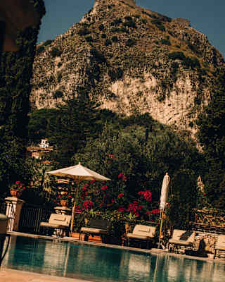 Ground-level view over the pool, where loungers recline beneath two parasols, one folded, with a dramatic mountain backdrop.