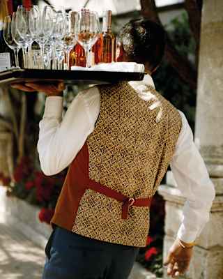 Barman in a gold waistcoat strolling away with a tray of wine and glasses on his shoulder