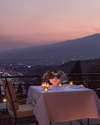 The silhouette of Mount Etna smoulders against the dusky pink of a sunset sky. Candle lanterns hang from a restaurant balcony