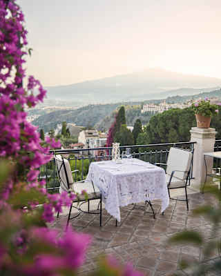 Bougainvillea tumbles down, framing the view of a lace topped terrace dining table for two, with Mount Etna rising behind