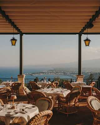 Laid tables with wicker chairs await guests on the Ristorante Timeo terrace, with a canopy and breathtaking coastline views.