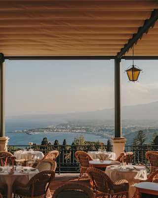 View across a covered restaurant terrace with tables and wicker chairs and extensive views over the stunning Ionian coast.
